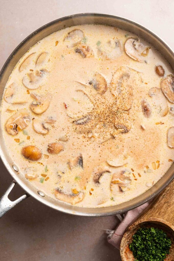 A skillet filled with creamy mushroom soup, reminiscent of the flavors in Cheesy Chicken Spaghetti Casserole, is garnished with ground pepper and sits on a light surface next to a small bowl of chopped herbs.