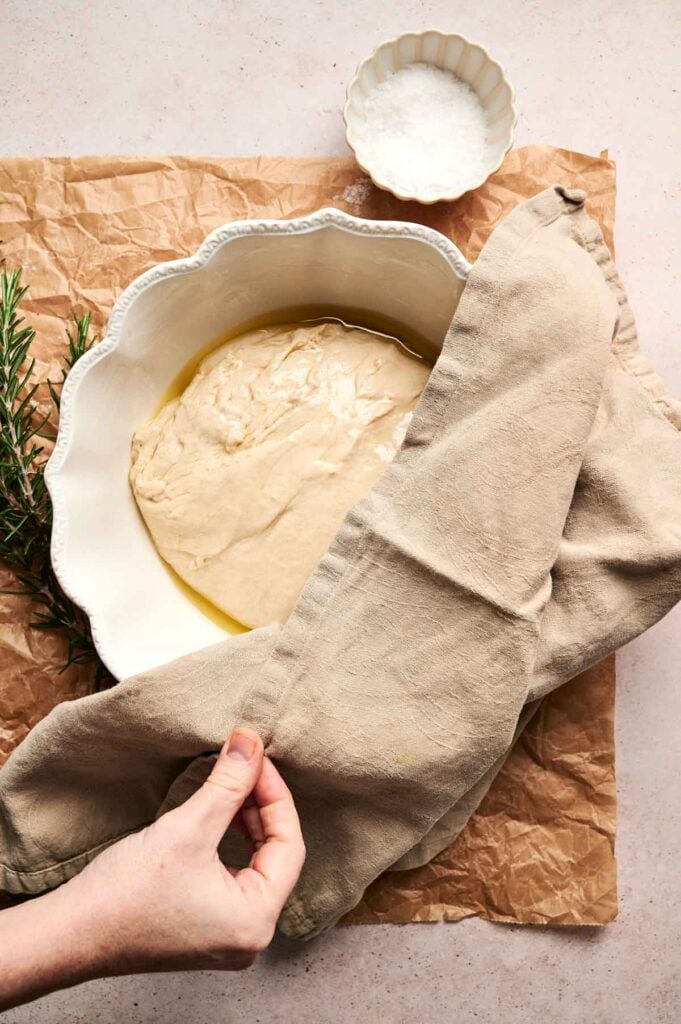A hand lifts a cloth covering a bowl of Air Fryer Focaccia Bread dough resting in oil, with a small bowl of salt and a rosemary sprig nearby on brown parchment paper.