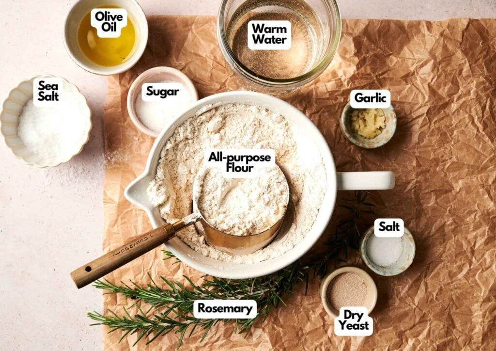 Ingredients for Air Fryer Focaccia Bread are arranged on a brown paper surface, including all-purpose flour, rosemary, sugar, salt, sea salt, dry yeast, garlic, olive oil, and warm water.