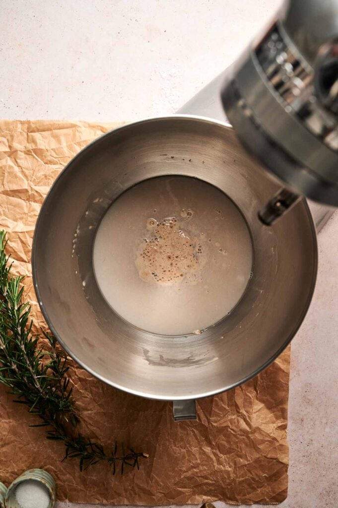 A mixing bowl with frothy yeast mixture inside sits on brown parchment paper, next to a sprig of rosemary&mdash;perfect for starting your Air Fryer Focaccia Bread&mdash;while a stand mixer attachment hovers overhead.