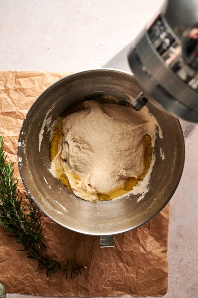 A stand mixer bowl containing Focaccia Bread dough with olive oil on the surface sits on parchment paper next to a sprig of fresh rosemary.
