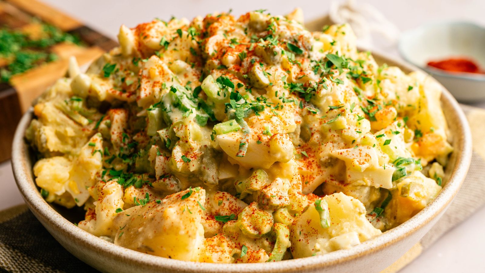 A bowl of potato salad topped with chopped herbs and a sprinkle of paprika, with visible chunks of potatoes and vegetables.