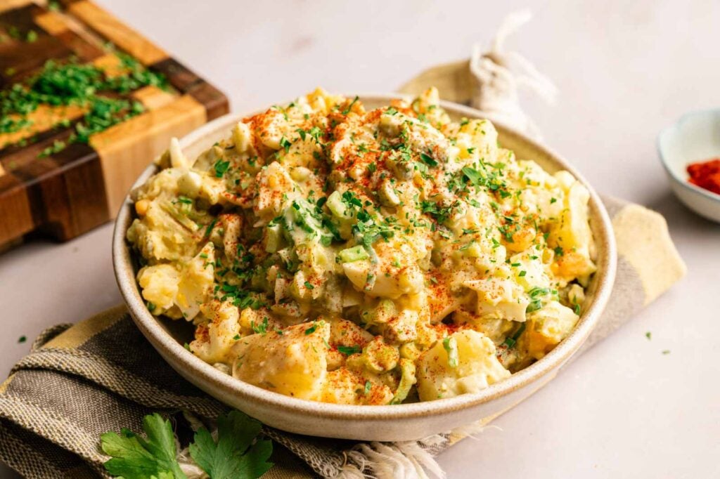 A bowl of Southern Potato Salad garnished with chopped herbs and paprika, placed on a striped cloth with a wooden cutting board and parsley in the background.
