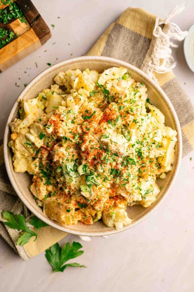 A bowl of Southern Potato Salad topped with chopped herbs and paprika sits on a striped cloth napkin, with fresh parsley nearby.