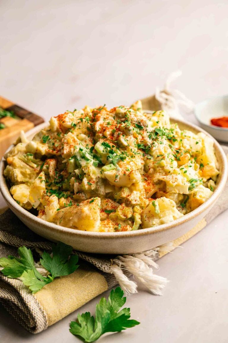A bowl of Southern Potato Salad garnished with chopped parsley and paprika, placed on a cloth napkin with fresh parsley on the side.