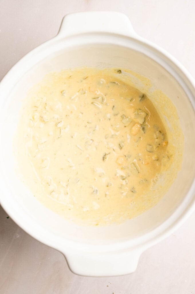 A white mixing bowl containing a thick, pale yellow dressing with visible chunks of vegetables, reminiscent of Southern Potato Salad dressing.