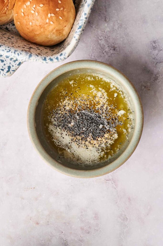 A bowl of melted butter topped with poppy seeds sits beside a tray of Air Fryer Turkey Sliders nestled in sesame seed buns on a light marble surface.