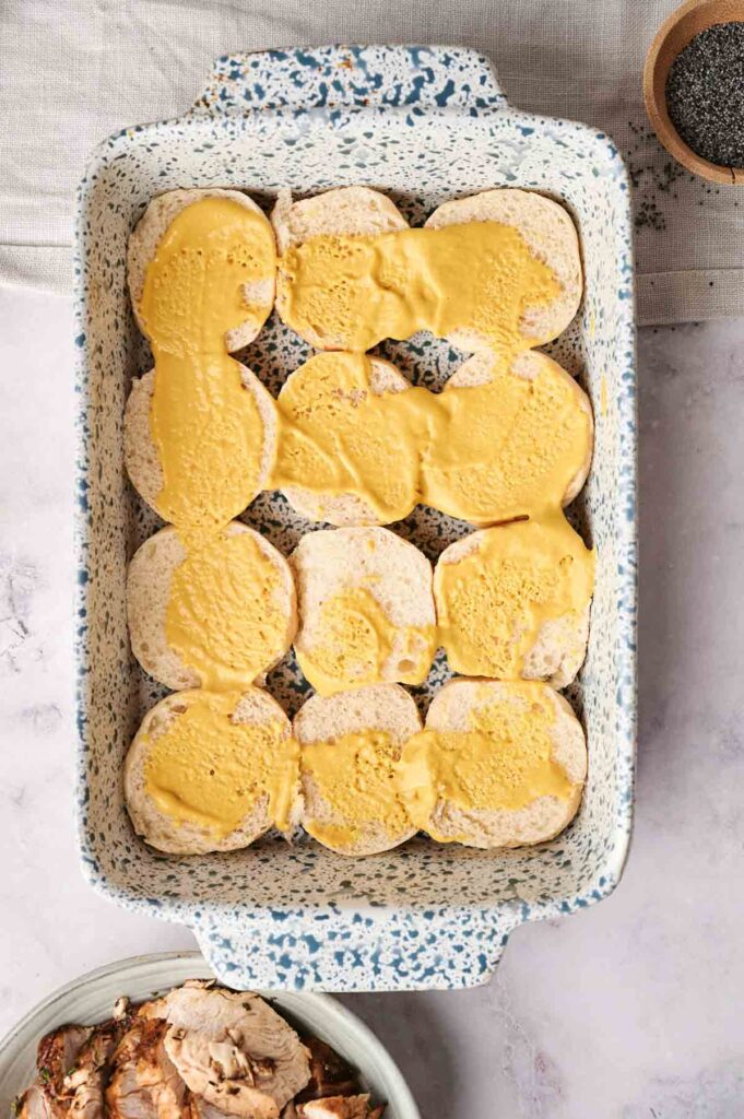 Rectangular speckled baking dish filled with sliced rolls partially covered in a yellow sauce, on a light surface, perfect for serving alongside Air Fryer Turkey Sliders; a small bowl of poppy seeds sits nearby.